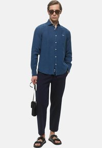 Camicia navy blue a maniche lunghe, con bottoni, e un logo bianco. Abbinata a pantaloni scuri e sandali neri; tiene in mano una borsa nera.