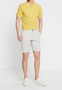 Pánské žluté polo tričko s bílým lemem, v kombinaci se světle šedými šortkami a bílými teniskami, stojící na jednolitém pozadí.