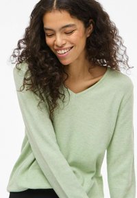 Jeune femme aux cheveux bouclés et foncés souriant doucement, portant un pull vert clair à col en V sur un fond blanc uni.