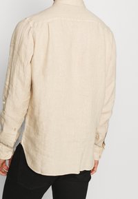 Camicia in lino beige con una vestibilità rilassata, maniche lunghe, cuspidature sui polsini e spacco laterale per maggiore comfort. Tessuto strutturato visibile.