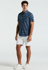 Marineblauw kortekorte mouw overhemd met witte palmbladprint, enkele borstzak, gedragen met witte shorts en lichte sneakers.