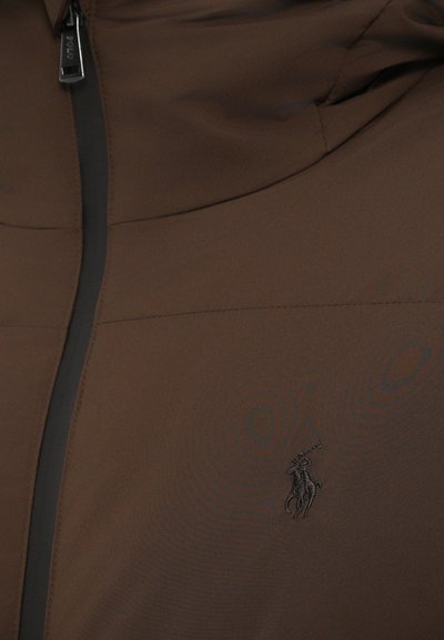 Hnedá zipsová bunda vyrobená z hladkej, ľahkej tkaniny. Obsahuje čierny zips a malé vyšívané logo na hornej ľavej strane.