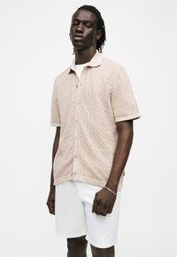 Άνδρας με σκουρόχρωμο δέρμα που φοράει μπεζ υφασμένο κοντομάνικο πουκάμισο με κουμπιά και λευκά σορτς σε μονόχρωμο λευκό φόντο.