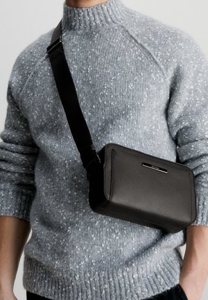 Czarna skórzana torba na ramię o prostokątnym kształcie, z srebrnym logo i czarnym regulowanym paskiem, noszona na szarym melanżowym swetrze.