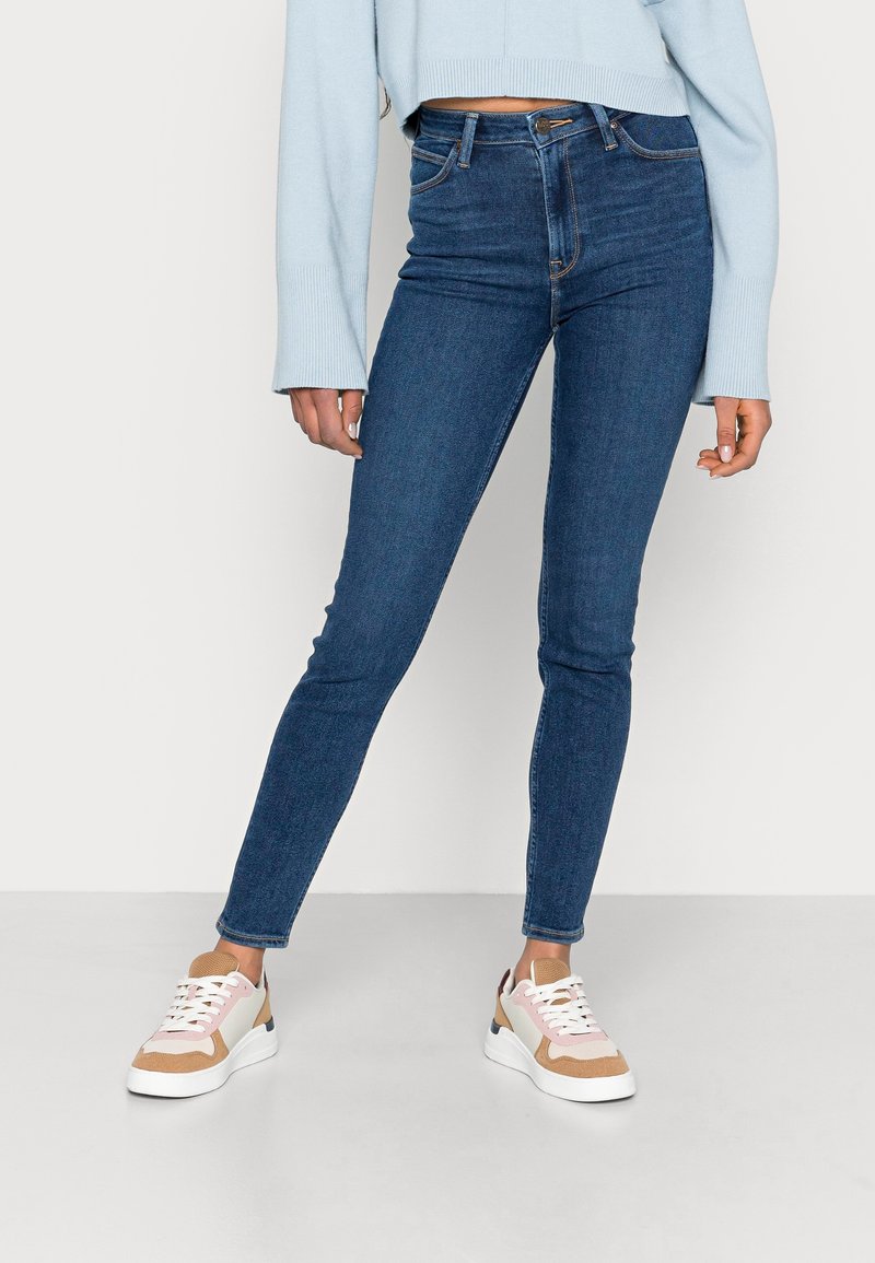 Jeans skinny de mezclilla azul con una cintura alta, que presentan una textura suave y zapatillas en tonos marrón y blanco con diferentes materiales y colores.