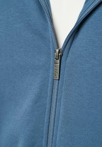 Blauwe ritsjack met een gladde textuur. Beschikt over een metallic rits met een label "USPA." De stof heeft een lichte glans en een uniform patroon.