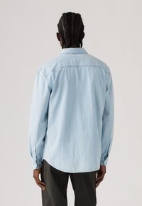 Cămașă din denim de culoare albastru deschis, cu mâneci lungi, guler tip buton și tiv drept. Prezintă cusături vizibile și manșete cu nasturi.