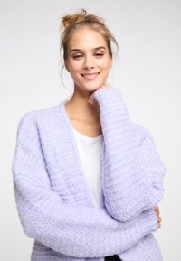 Ljust lila, fluffig stickad cardigan med ett texturerat mönster, öppen framsida och ribbade manschetter. Bärs över en vit topp och ger en avslappnad passform.