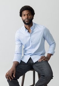 Svijetloplava košulja na dugim rukavima, s preklopljenim manžetama. Tamne traperice, sjedeći na drvenoj klupi. Neutralna pozadina.