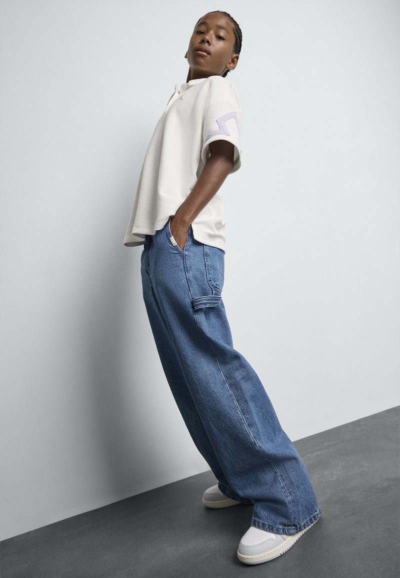 Polo blanco de manga corta con acentos morados, combinado con pantalones de mezclilla azul sueltos y zapatillas blancas, sobre un fondo gris claro.