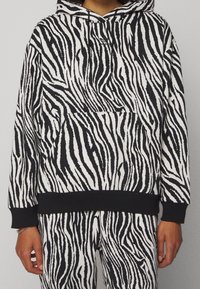 Sudadera de rayas de cebra en blanco y negro con un bolsillo tipo canguro, capucha con cordón y puños y dobladillo de canalé negros. Textura de tela suave.