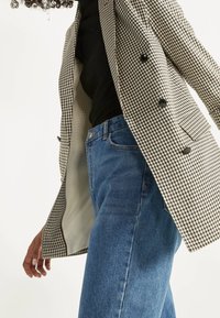 Dubbelgeknoopte geruite blazer in bruin en wit, gecombineerd met blauwe spijkerbroek. Getextureerde stof en zwarte knoopdetails zijn zichtbaar.