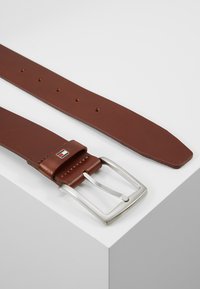 Ceinture en cuir marron avec une boucle en métal argenté, dotée de plusieurs trous pour l'ajustement et d'un accent de marque près de la boucle.