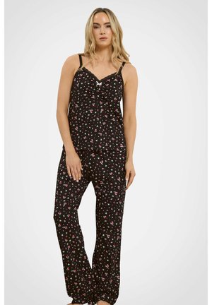 Donna che indossa una canottiera nera a fiori e pantaloni abbinati, in piedi davanti a uno sfondo chiaro e uniforme.