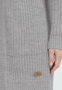 Cardigan de punto gris con un patrón texturizado, con puños y dobladillo acanalados, y una pequeña etiqueta de cuero con "oxmo" en el costado.