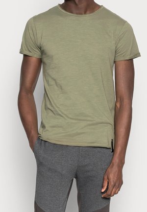 Hombre vistiendo una camiseta de manga corta verde oliva y pantalones gris oscuro, de pie con una mano en el bolsillo contra un fondo blanco.