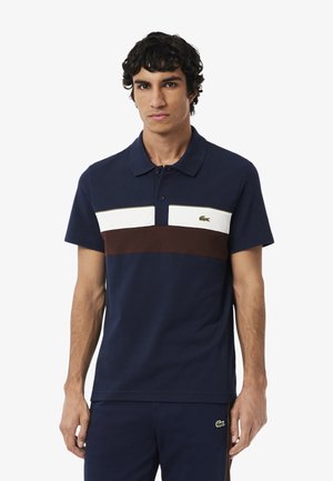 Polo navy con righe orizzontali bianche e marroni, dotato di un colletto classico e del logo verde Lacoste sul petto. Materiale in cotone.