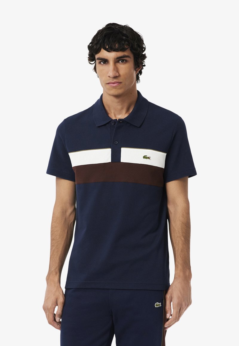 Granatowa koszulka polo w białe i brązowe poziome paski, z klasycznym kołnierzykiem i zielonym logo Lacoste na piersi. Wykonana z bawełny.