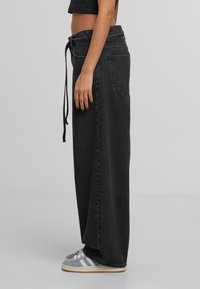 Sivuprofiili henkilöstä, joka on pukeutunut pitkä musta farkuhame, jossa on solmittava vyötärö ja harmaat lenkkarit valkoisilla raidoilla harmaata taustaa vasten.