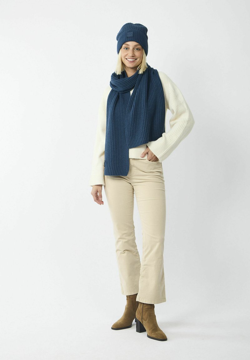 Gebreide blauwe beanie en sjaal, crème trui, beige broek en bruine gehakte enkellaarsjes. De sjaal heeft een gestructureerd ribpatroon.