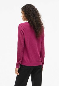 Maglione fucsia a maglia con scollatura rotonda e orlo a costine. La modella lo indossa con pantaloni neri, mostrando i capelli ricci di spalle.