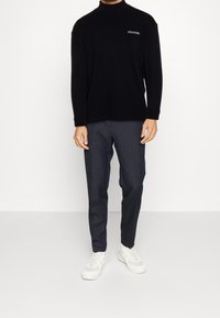 Mies, który ma na sobie czarny top z długim rękawem z napisem "HILFIGER", ciemnoszare spodnie o kroju slim oraz białe sneakersy, stojący na tle jednolitym.