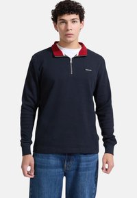 Maglione blu navy a coste con colletto rosso e zip a quarto, con etichetta logo. Indossato con jeans blu, tessuto a trama.