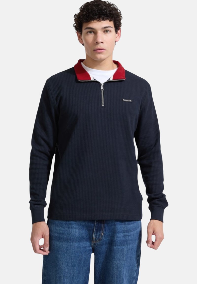 Maglione blu navy a coste con colletto rosso e zip a quarto, con etichetta logo. Indossato con jeans blu, tessuto a trama.