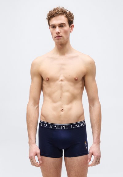 Νεαρός άνδρας με σγουρά μαλλιά, φορώντας boxer briefs Polo Ralph Lauren σε σκούρο μπλε, στέκεται μπροστά σε απλό ανοιχτό γκρι φόντο.