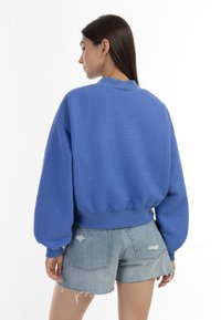 Felpa blu con texture, colletto rotondo e polsini a costine, abbinata a pantaloni corti di jeans strappati. La felpa ha una vestibilità ampia.