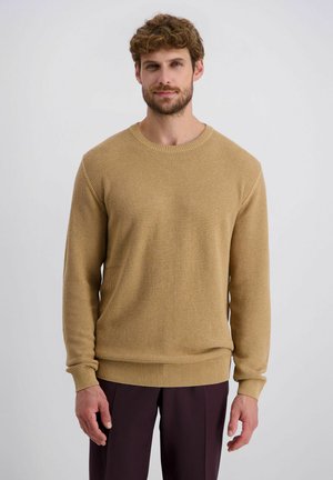 Man staand, gekleed in een beige gebreide trui met textuur en donkerbruine broek, tegen een effen lichte achtergrond.