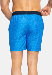 Pantaloni da bagno blu con pois bianchi, vita elastica e tasche laterali. Tessuto strutturato e dettaglio di una tasca posteriore.