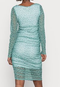 Robe teal froncée avec de longues manches transparentes, présentant un motif floral en blanc et rose. Texture lisse avec une silhouette ajustée.