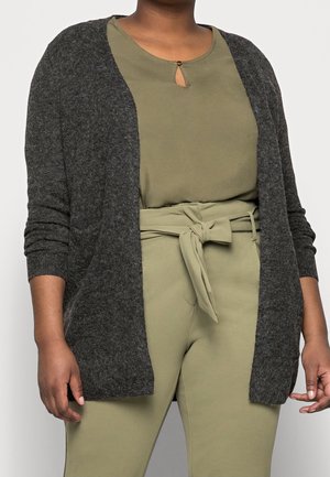 Persoon in olijfgroene broek met riem en bijpassende top met een kleine keyhole halslijn, gelaagd met een donkergrijze open cardigan.