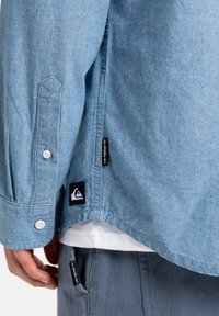 Camisa de denim azul con mangas largas, puños abotonados y un dobladillo curvado. Presenta una etiqueta de logo en el costado y costuras en blanco en contraste.