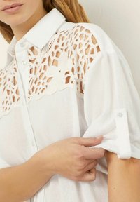 Blusa bianca con colletto abbottonato, dettagli in pizzo sulle spalle e maniche arrotolate, realizzata in tessuto leggero e testurizzato.