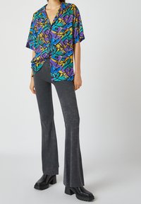 Camisa de manga corta en un vibrante patrón geométrico de púrpura, verde y amarillo; combinada con pantalones acampanados gris oscuro y botas negras gruesas.
