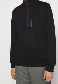 Sweatshirt noir avec un col montant, arborant une fermeture éclair colorée à moitié et une petite poche avant. Tissu doux avec un ourlet côtelé.