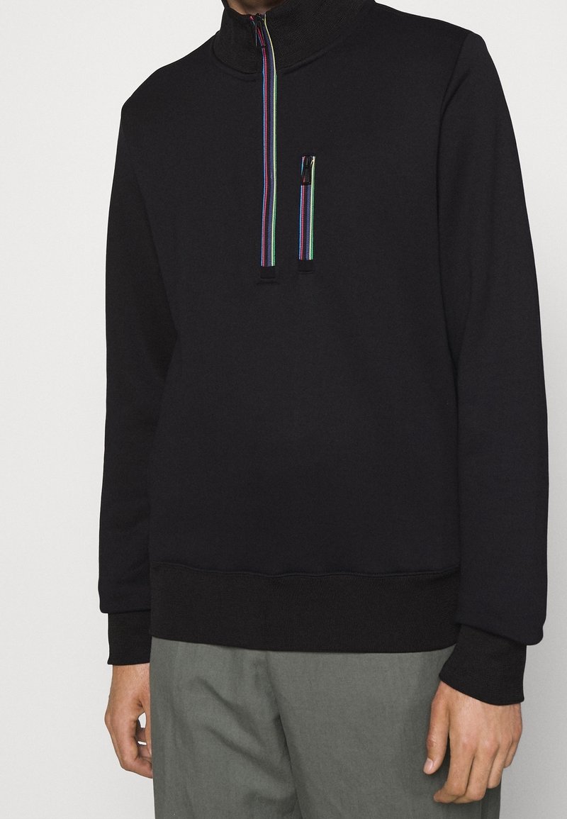 Sweatshirt noir avec un col montant, arborant une fermeture éclair colorée à moitié et une petite poche avant. Tissu doux avec un ourlet côtelé.