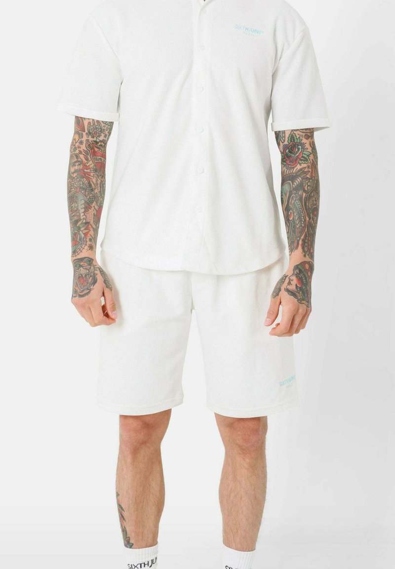 Chemise blanche à manches courtes, à boutons, avec un motif texturé, assortie à un short blanc. Comprend un détail de logo bleu clair.
