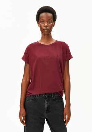 Frau mit kurzem lockigem Haar, die ein burgunderfarbenes Kurzarm-T-Shirt und schwarze hoch taillierte Jeans vor einem einfarbigen hellen Hintergrund trägt.