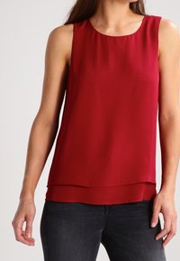 Blouse rouge sans manches à couches portée avec un jean gris foncé, montrée sur une personne debout contre un fond blanc uni.