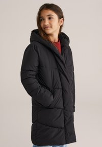 Jauna meitene, tērpta garā melnā puffera jakā ar kapuci, rokas kabatās, skatās nedaudz no sāna uz vienkārša fona.