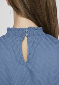 Niebieska bluzka o teksturowej powierzchni z marszczonym kołnierzykiem, z otworem w kształcie łezki z tyłu i dwoma dekoracyjnymi guzikami. Widoczny wzór w zygzaki.