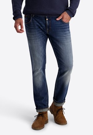 Mann trägt hochgekrempelte dunkelblaue Jeans, einen dunkelblauen strukturierten Pullover und braune Wildleder-Schnürstiefel, steht mit einer Hand in der Tasche.