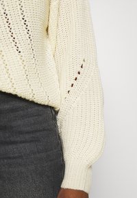 Strickpullover in Creme, mit lockerem Schnitt, Zickzackmustern und gerippten Bündchen. Trägt man über einem dunkelgrauen Stoff.