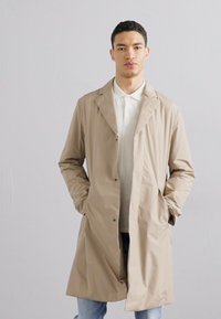 Beige lightweight coat with a collar, open front, and black buttons. Person wears a white shirt underneath with blue jeans. Solid color, smooth texture.