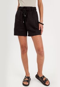 Shorts en lin noirs avec une taille élastique et un cordon de serrage, offrant une coupe décontractée. Associés à des sandales noires, mettant en avant un accent de boucle.