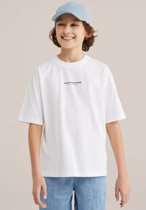 Smiling young person with curly hair wearing a light blue cap, white "Blue Ridge Denim" t-shirt, and light blue jeans against beige background.