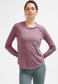 Langärmliges Sportoberteil in hellem Lila mit Rundhalsausschnitt, strukturierter Stoff und einem Logo auf der Brust. Kombiniert mit hochtaillierten grünen Leggings.
